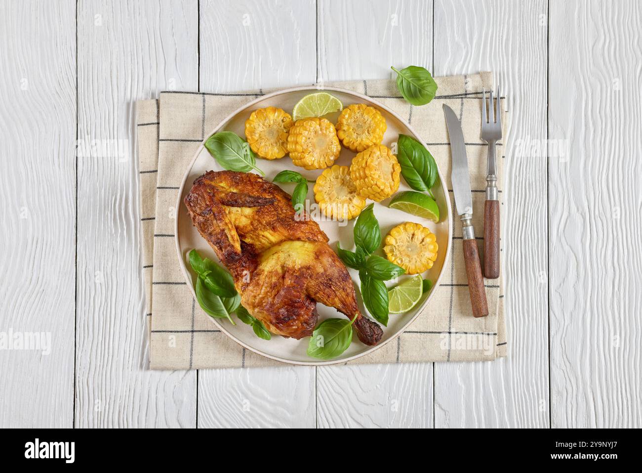 la moitié du poulet bbq avec des pièces d'épis de maïs et des feuilles de basilic frais sur l'assiette sur une table en bois blanc avec des couverts, vue horizontale d'en haut, pose à plat Banque D'Images
