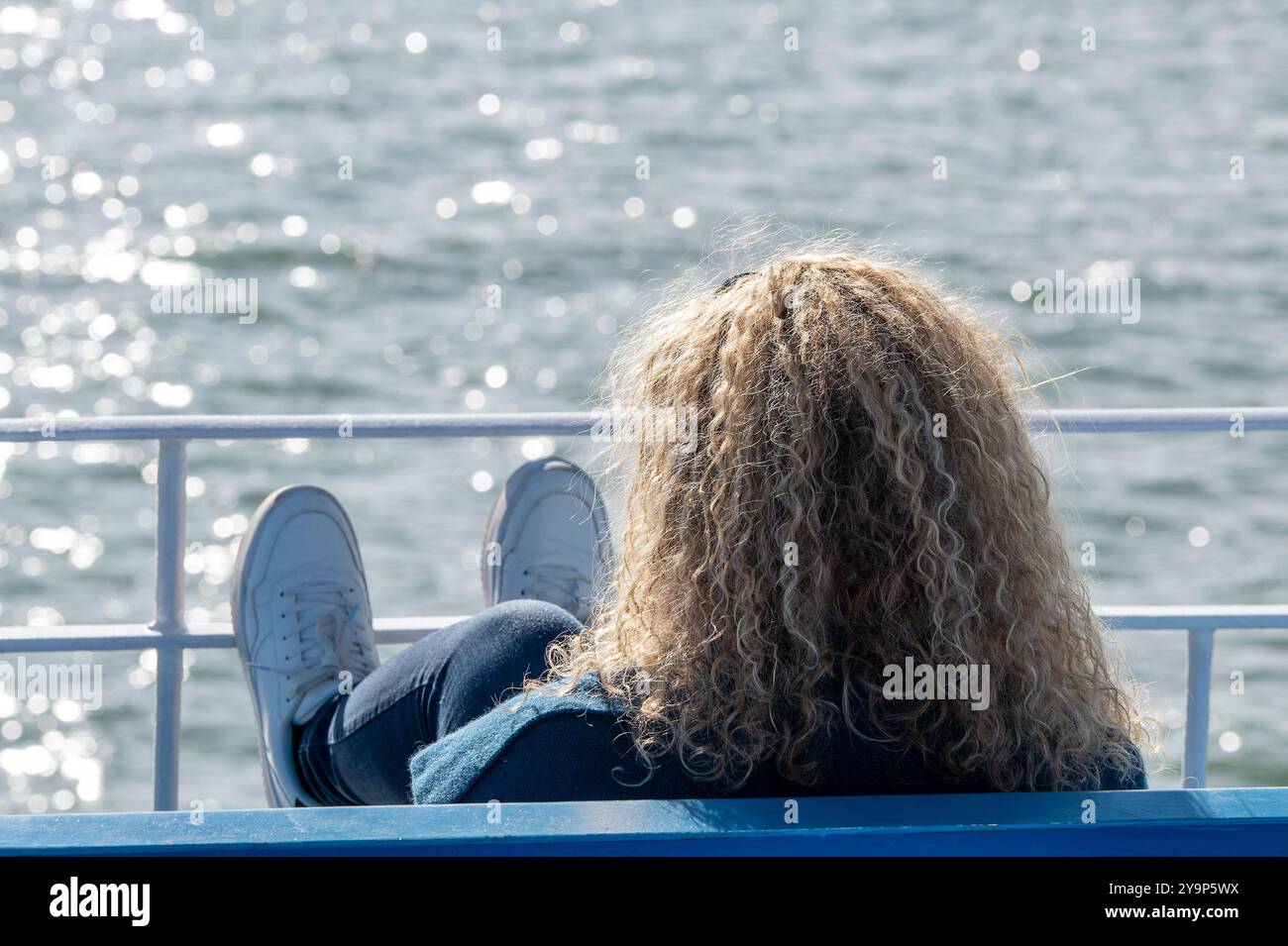 jeune femme aux cheveux permanents portant une veste en denim et des chaussures d'entraînement blanches assis avec ses pieds sur des rampes regardant vers la mer par une journée ensoleillée. Banque D'Images