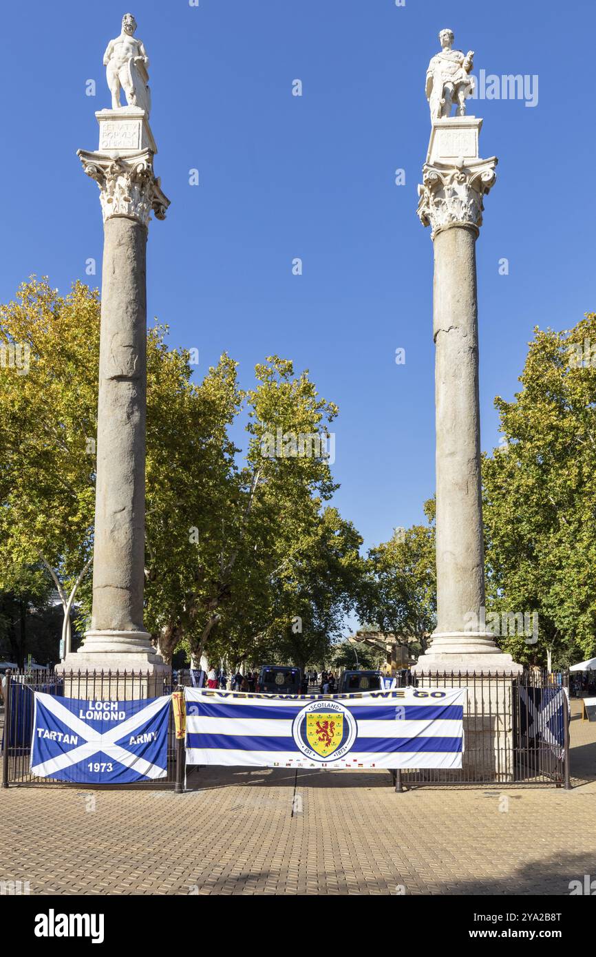 Deux imposantes colonnes avec des statues flanquées d'arbres et de drapeaux sous un ciel bleu vif, Séville Banque D'Images Deux imposantes colonnes avec des statues flanquées d'arbres et de drapeaux sous un ciel bleu vif, Séville Banque D'Images