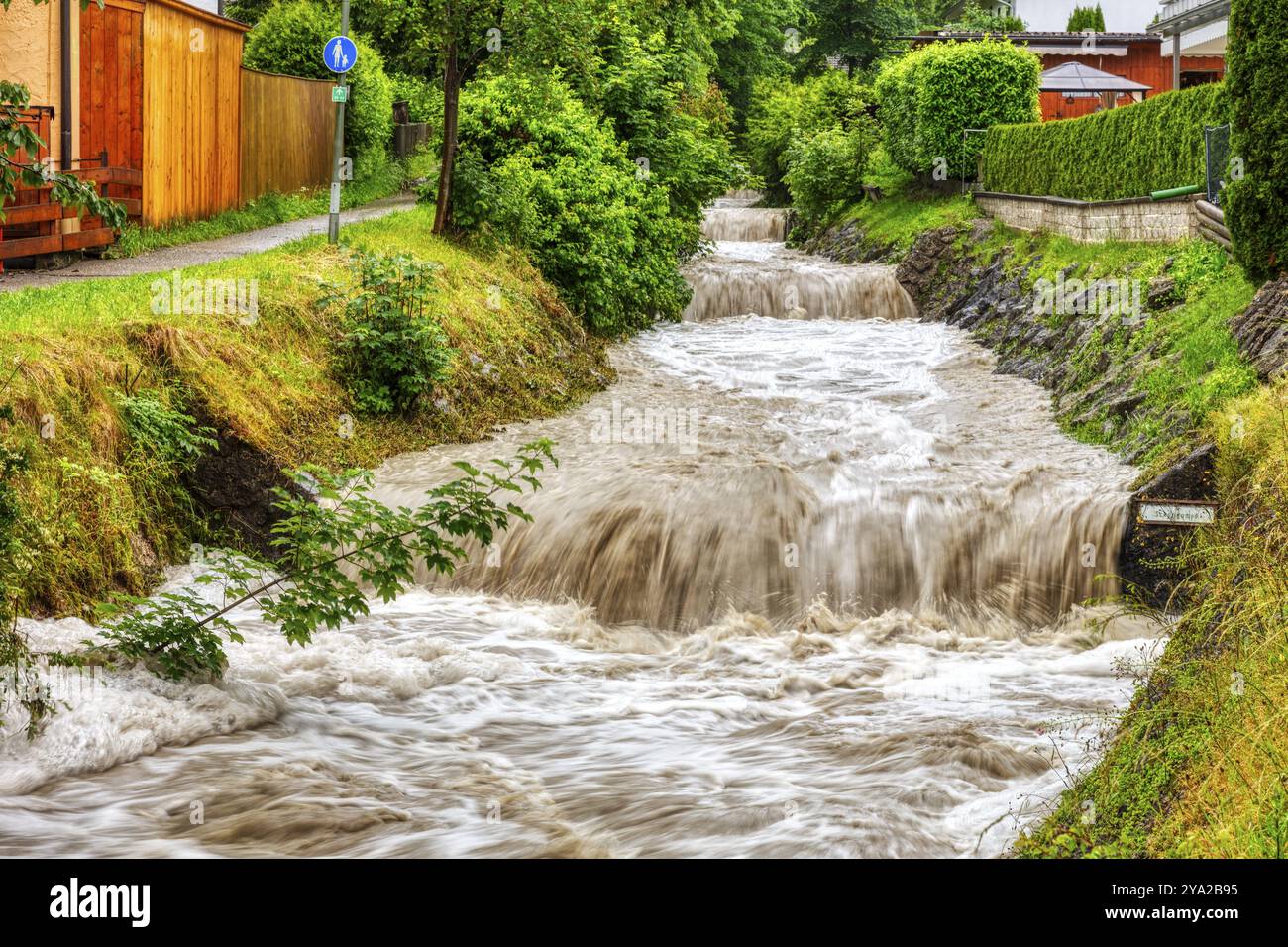 Ruisseau en forme d'inondation dans une zone de banlieue, encadrée par des arbres et des bâtiments modernes, Osterbach, Bad Feilnbach Banque D'Images Ruisseau en forme d'inondation dans une zone de banlieue, encadrée par des arbres et des bâtiments modernes, Osterbach, Bad Feilnbach Banque D'Images