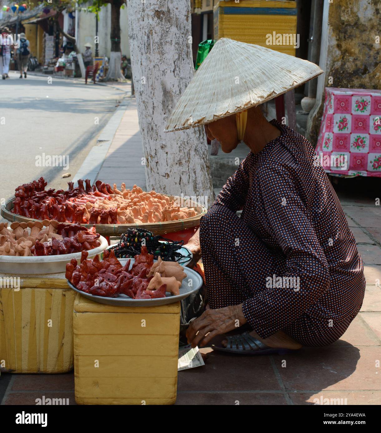 03/23/2017 - Hoi an, Vietnam : un vendeur de rue traditionnel vêtu d'un chapeau conique, expose une variété d'artisanat fait main le long d'une rue animée, illust Banque D'Images