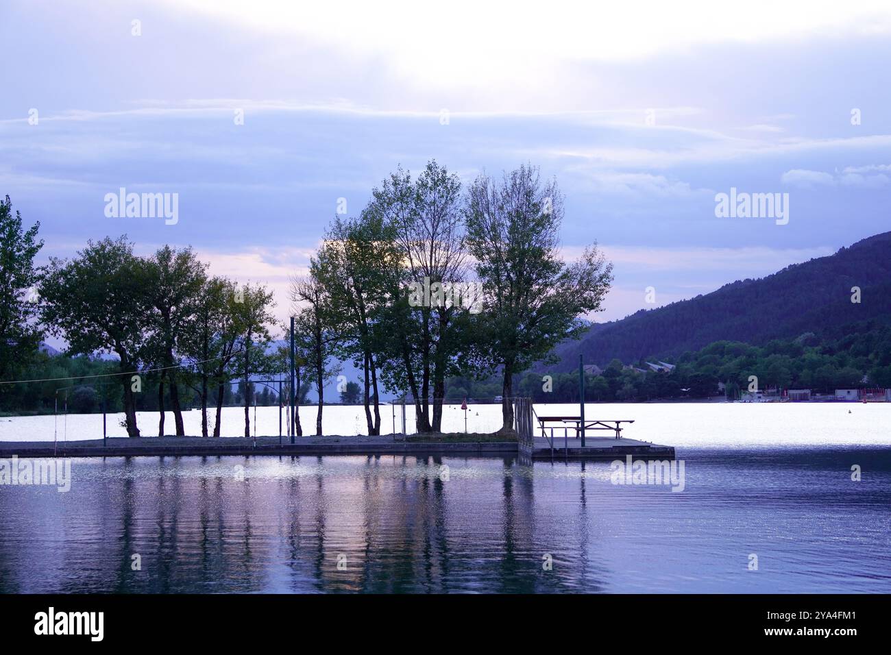 Scène paisible d'un lac au coucher du soleil avec des arbres reflétés sur l'eau, créant une atmosphère calme et sereine sous un ciel légèrement nuageux. Banque D'Images