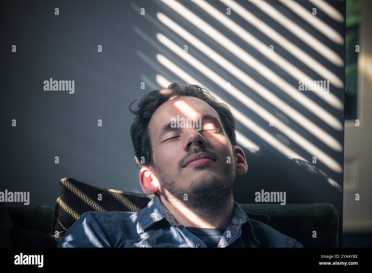 Portrait d'un homme caucasien somnolant sur le canapé, sur le visage tombe l'ombre des stores Banque D'Images