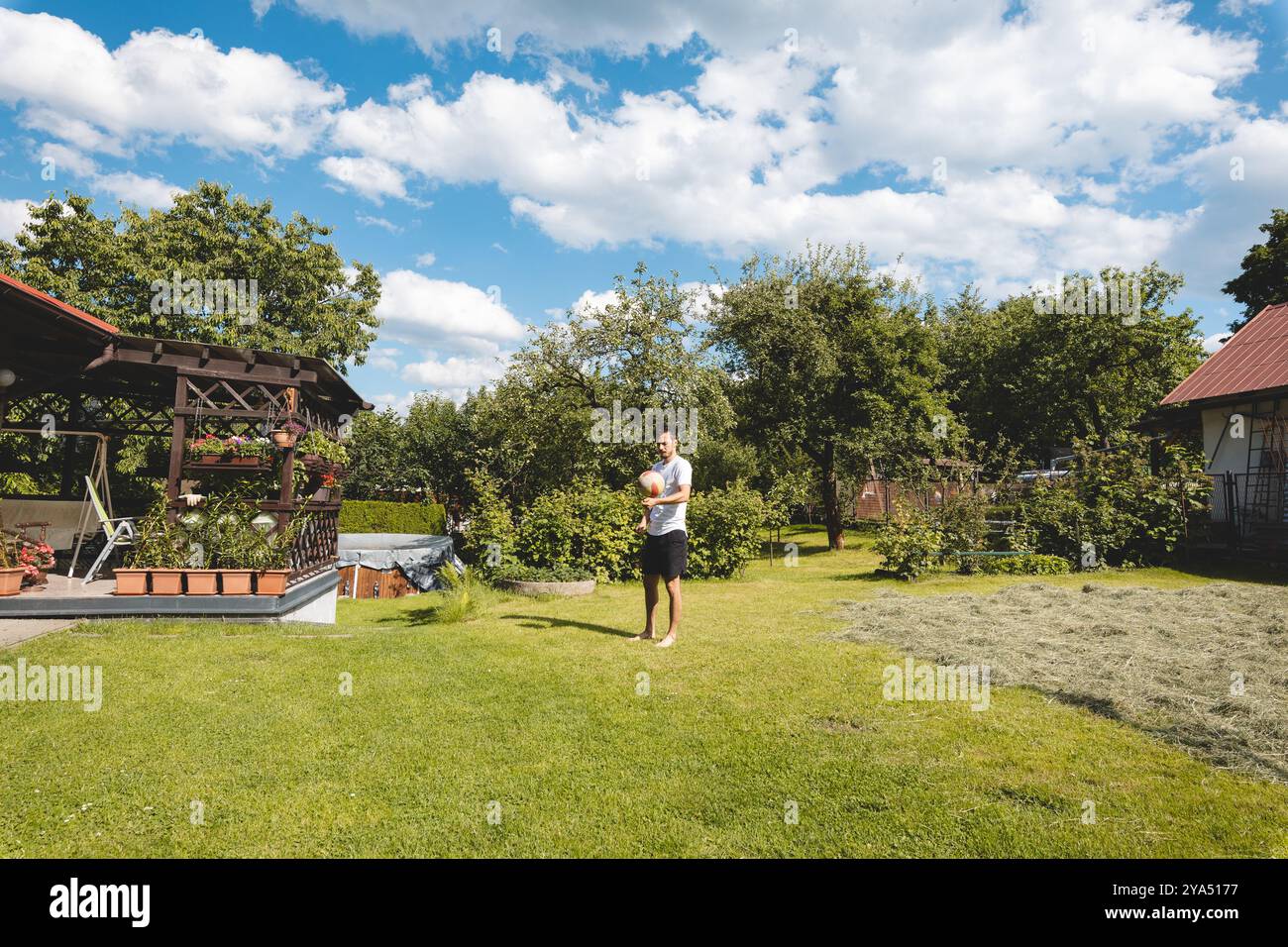 le joueur pratique une bosse de volley-ball dans un jardin plein d'arbres et d'herbe. Il est vêtu d'un t-shirt blanc et d'un short noir, pieds nus. Le ciel bleu avec Banque D'Images