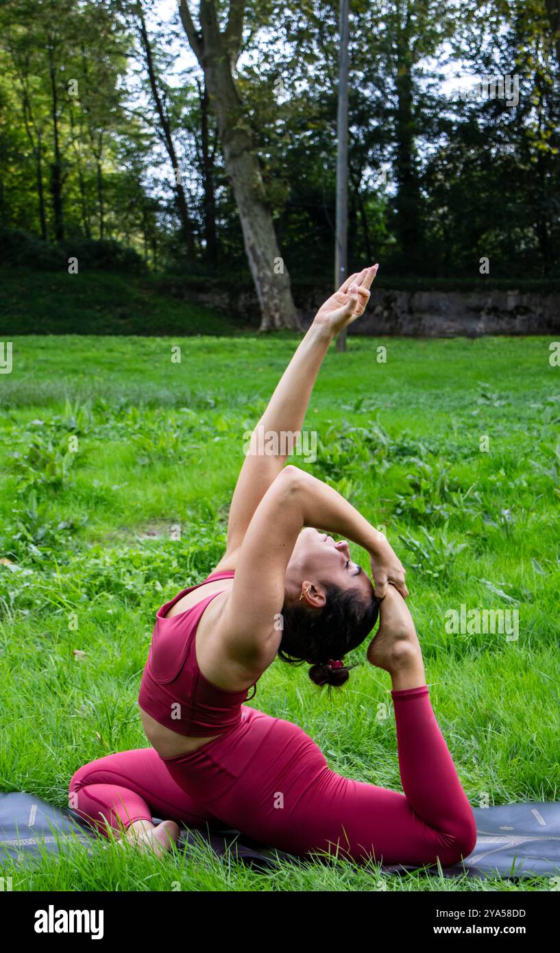 Plan vertical d'une belle jeune femme avec ses cheveux attachés en arrière exécutant une pose de yoga difficile sur son tapis sur l'herbe verte du parc reliant Banque D'Images