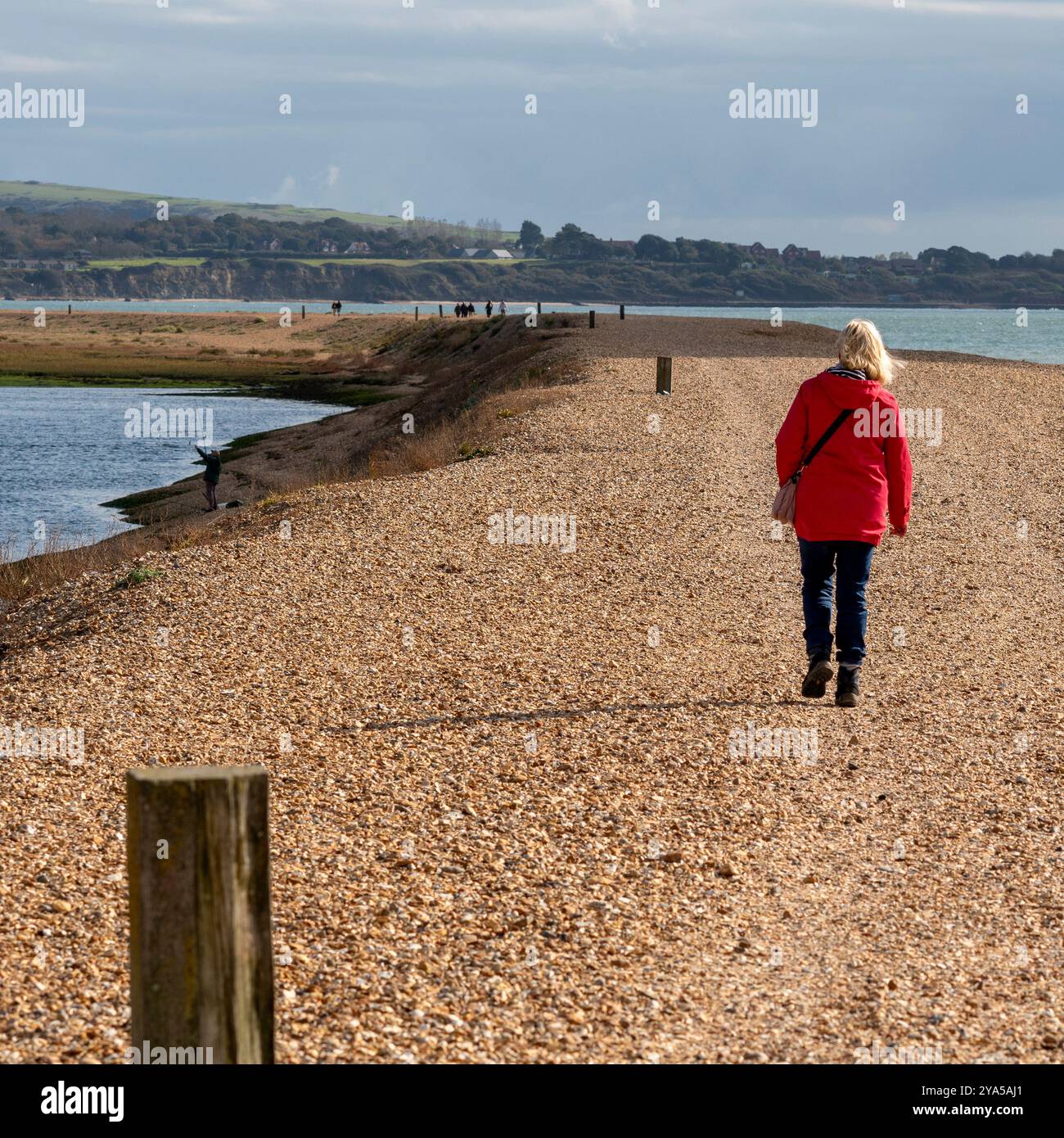 Femme en manteau rouge marchant sur la banque de galets Hurst Spit, Keyhaven, Hampshire, Angleterre, Royaume-Uni Banque D'Images