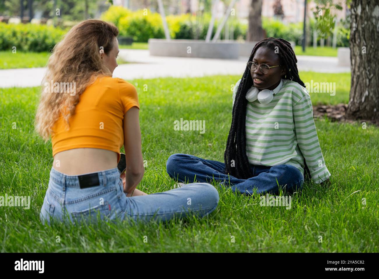 Deux jeunes femmes sont assises sur l'herbe dans un parc, profitant du temps estival et de l'autre compagnie pendant qu'elles s'engagent dans la conversation Banque D'Images