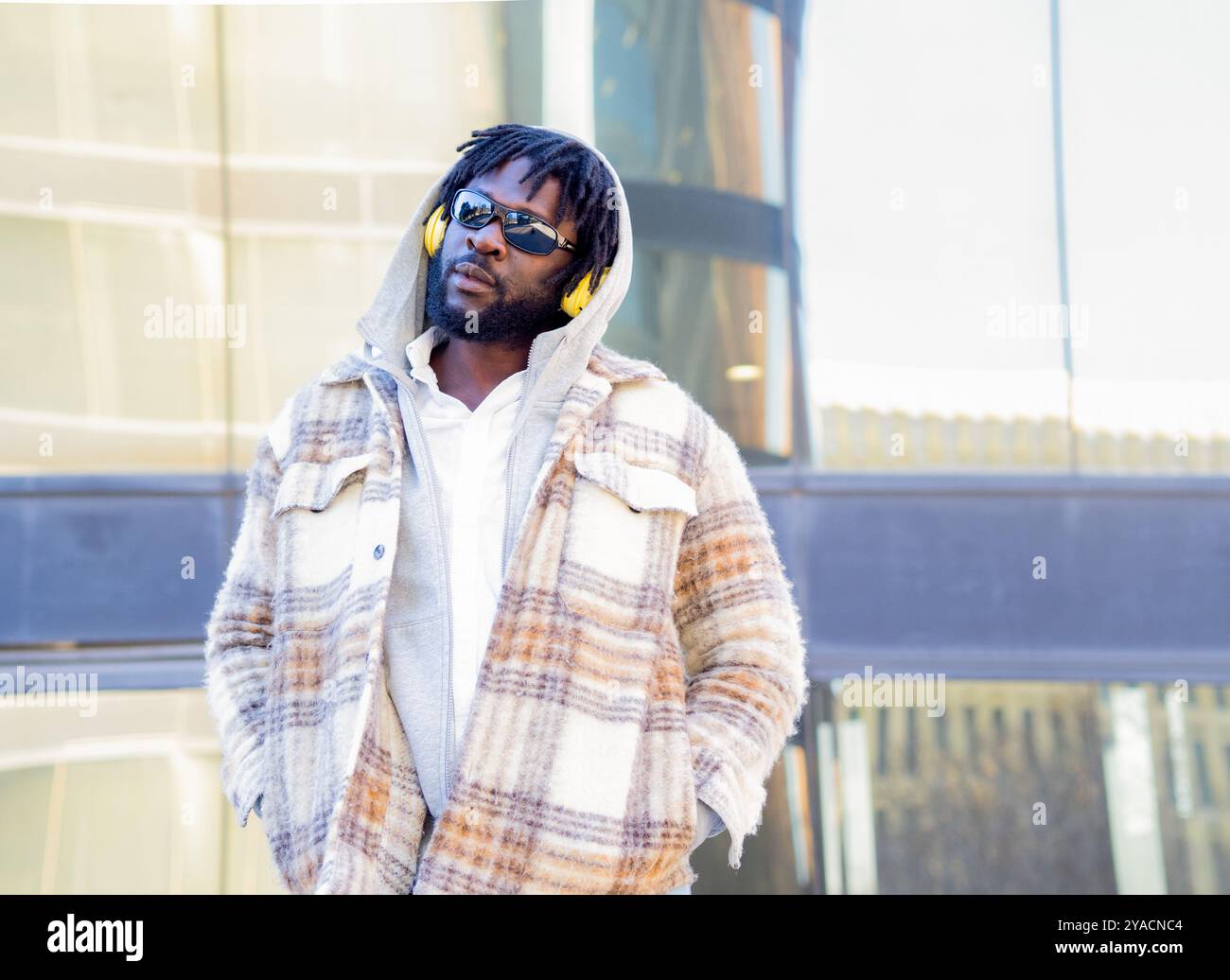 Portrait d'un homme africain avec des lunettes de soleil, des dreadlocks et des écouteurs regardant la caméra dans une ville. Banque D'Images