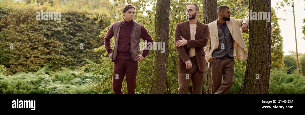 Vêtus d'une tenue d'automne chic, trois hommes se tiennent confiants dans un champ coloré entouré par la nature. Banque D'Images