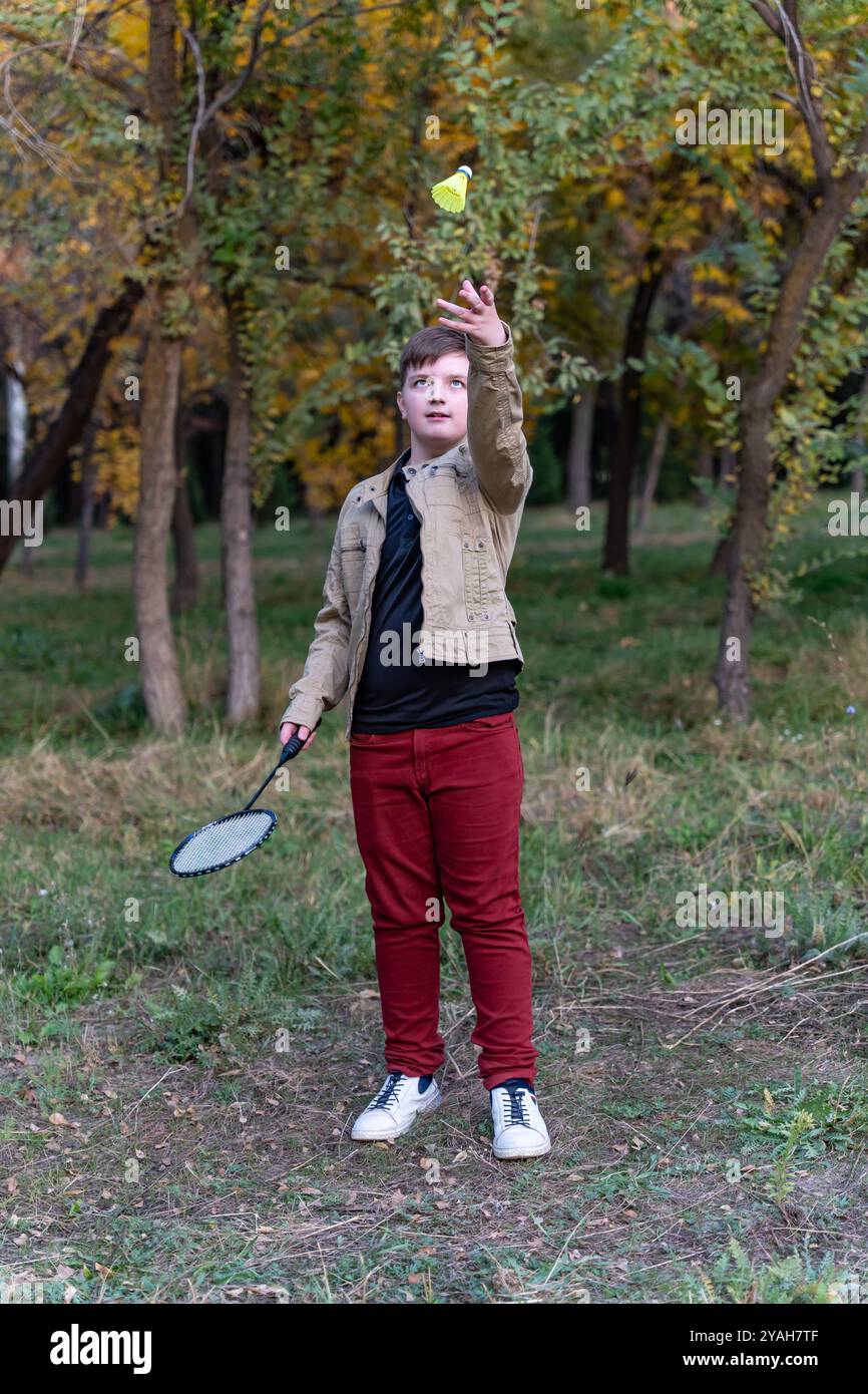 Un adolescent en veste beige, Jean Bordeaux et baskets blanches se tient dans un parc d'automne, tenant une raquette de badminton dans sa main droite. Banque D'Images