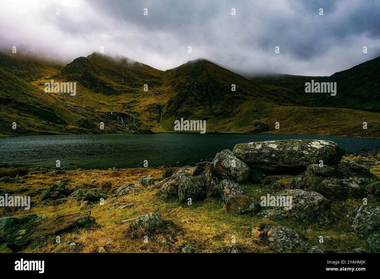 Un lac calme et serein entouré de collines verdoyantes. De grands rochers jonchent le premier plan, tandis qu'une fine cascade ruisselle le long de la montagne. Banque D'Images