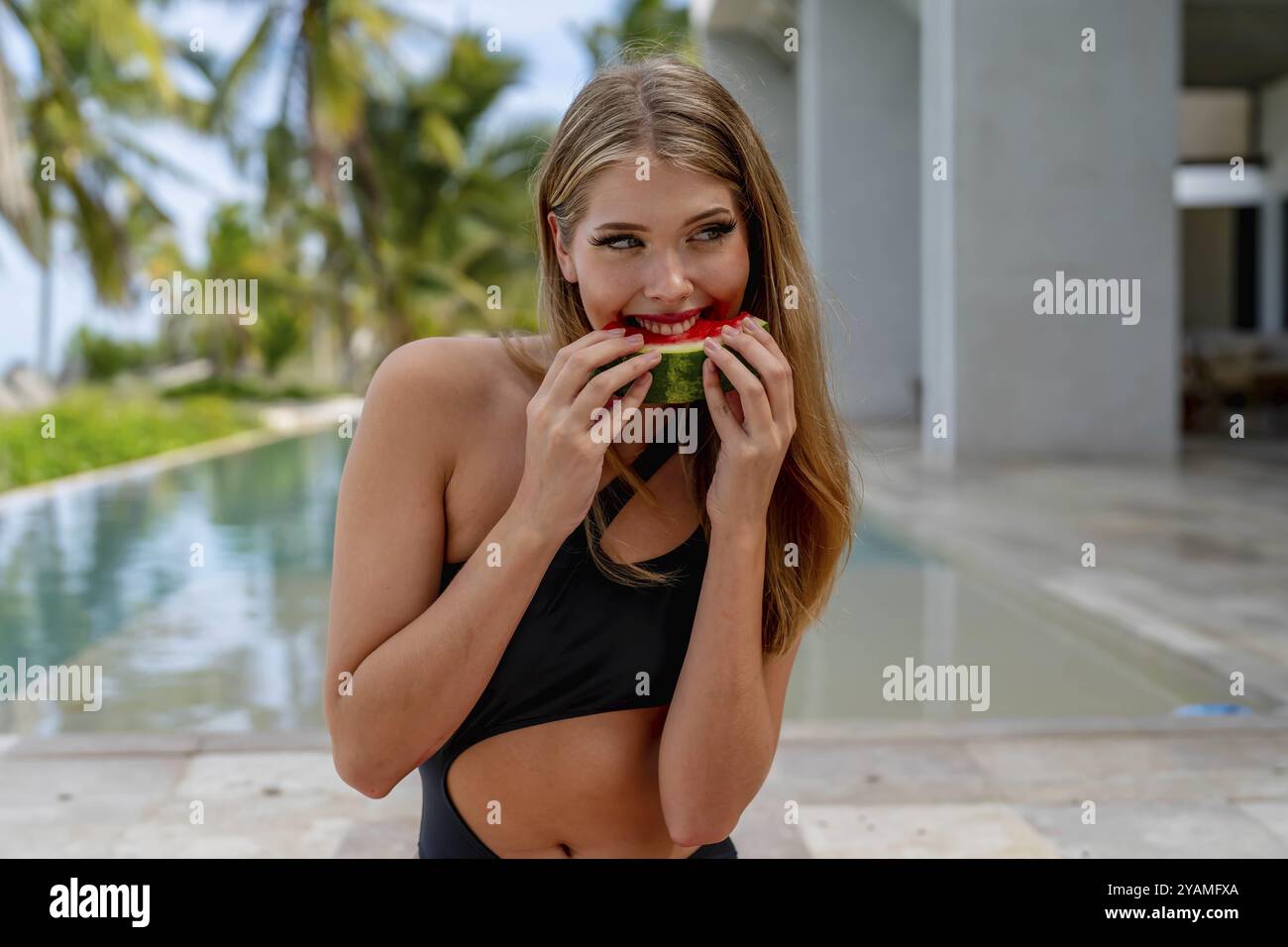 Rayonnant au bord de la piscine, une jeune beauté se prélasse dans le bonheur des vacances. Son sourire scintille tandis qu'elle dévore savamment la pastèque, incarnant l'allu estival insouciant Banque D'Images