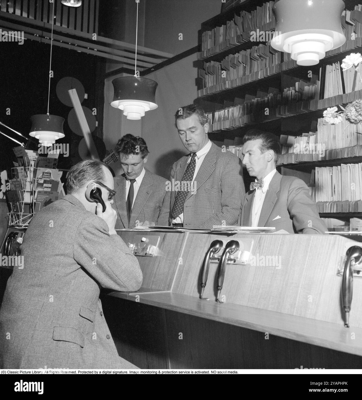 Comment vous avez acheté des disques dans les années 1950 L'homme tenant ce qui ressemble à un téléphone écoute en fait un disque. À cette époque, il était courant que les clients puissent écouter les disques dans le magasin avant de décider de les acheter. Le magasin de disques dispose de plusieurs stations d'écoute et le personnel derrière le comptoir gère le système. Ce magasin particulier est Selanders skivbar sur Vasagatan à Stockholm. 1955. Banque D'Images