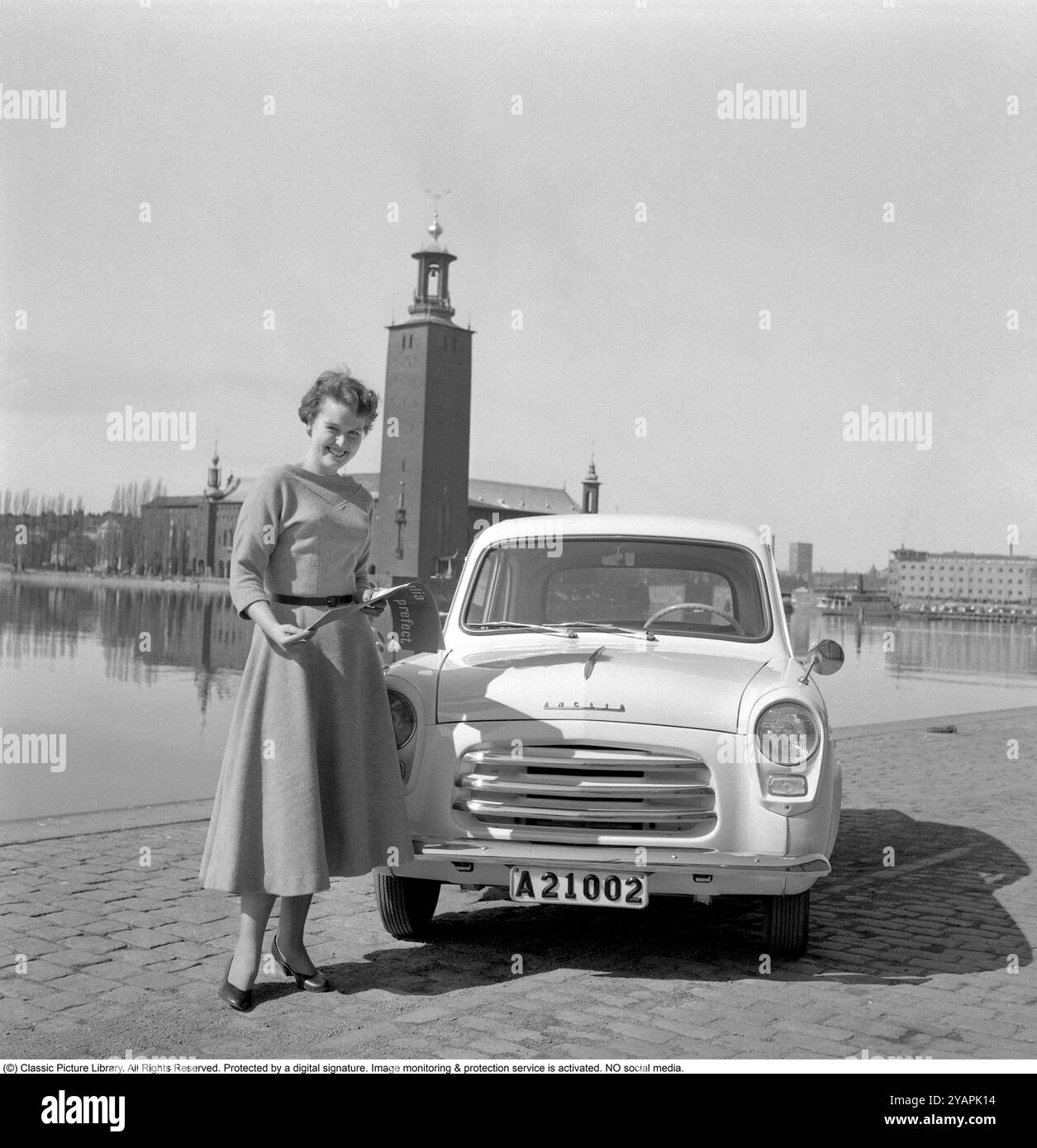 Voiture Ford dans les années 1950 Une jeune femme à côté d'une voiture Ford Anglia dans le centre de Stockholm avec l'hôtel de ville en arrière-plan. 1956 Banque D'Images
