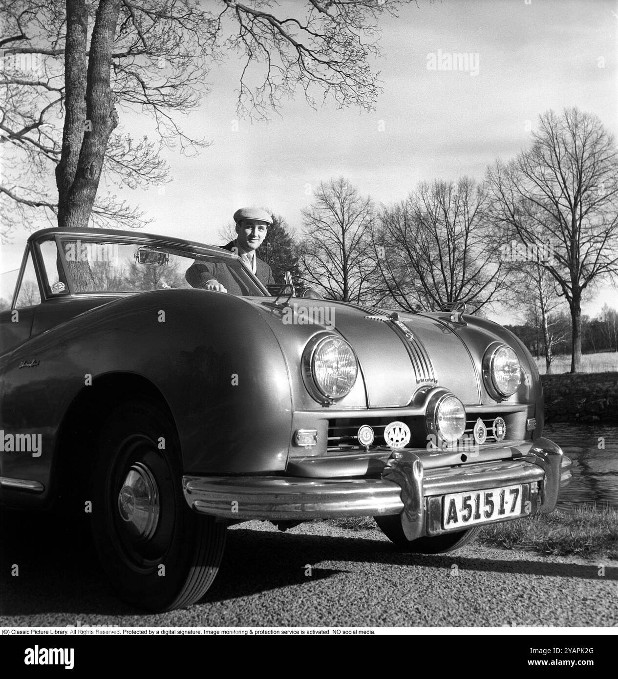 Un fier propriétaire de voiture dans les années 1950 Un homme dans sa voiture, une Austin A 90 Atlantic de 1950 avec le numéro d'immatriculation A51517. La voiture a été fabriquée par le constructeur automobile britannique Austin. Le modèle était initialement destiné à être vendu sur le marché américain. Il avait un corps ouvert, un cabriolet. La voiture avait un moteur à essence 4 cylindres, 4 vitesses et une propulsion arrière. La voiture avait trois phares. Ce propriétaire de voiture était membre de plusieurs associations automobiles internationales et leurs emblèmes sont montés sur le devant de la voiture. Anders Svahn ref SVA13 Banque D'Images