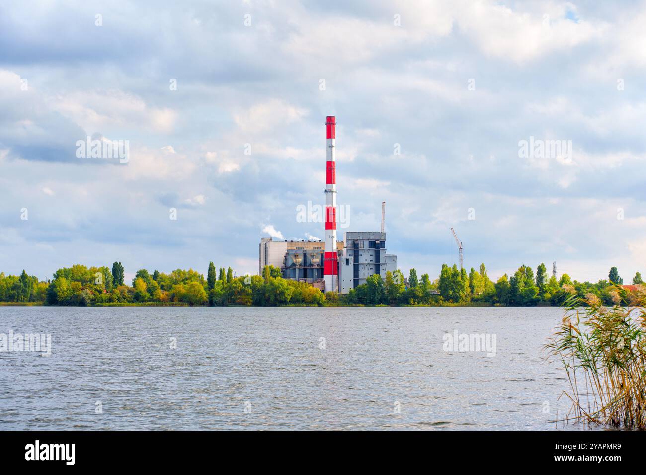 Usine d'incinération de déchets située près d'un lac serein, entourée d'arbres sous un ciel nuageux. Banque D'Images