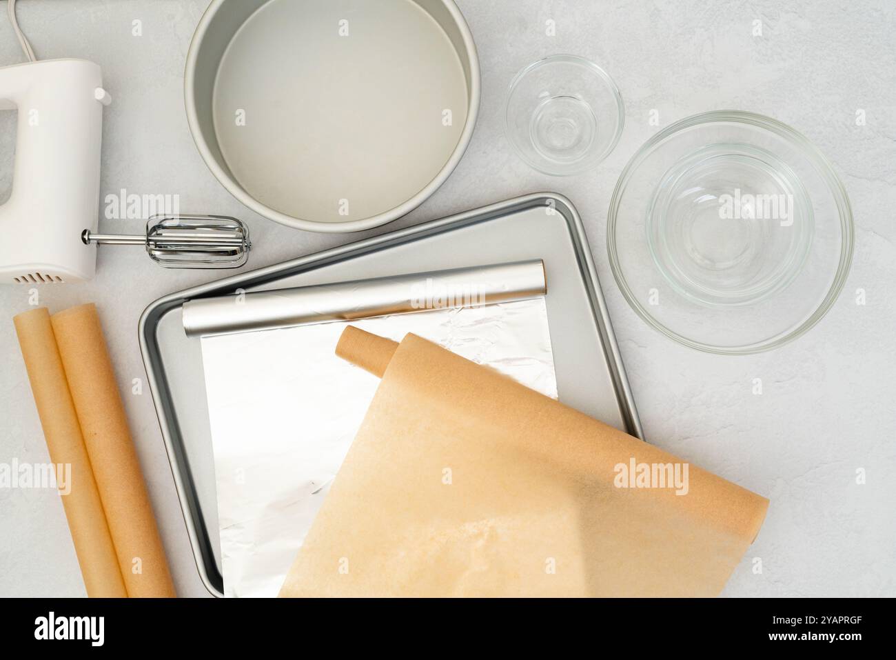 Composition à poser à plat comprenant du papier parchemin, une feuille d'aluminium, un moule à pâtisserie, un mélangeur et des bols en verre disposés en vue rapprochée sur un dos gris clair Banque D'Images