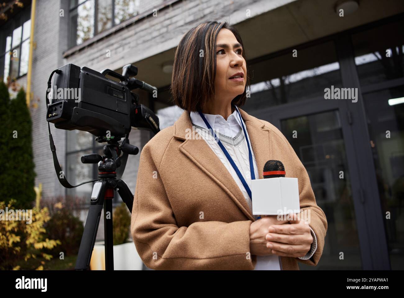 Une journaliste multiraciale vêtue d'un manteau beige se tient confiante à l'extérieur, se préparant à rapporter des nouvelles. Banque D'Images