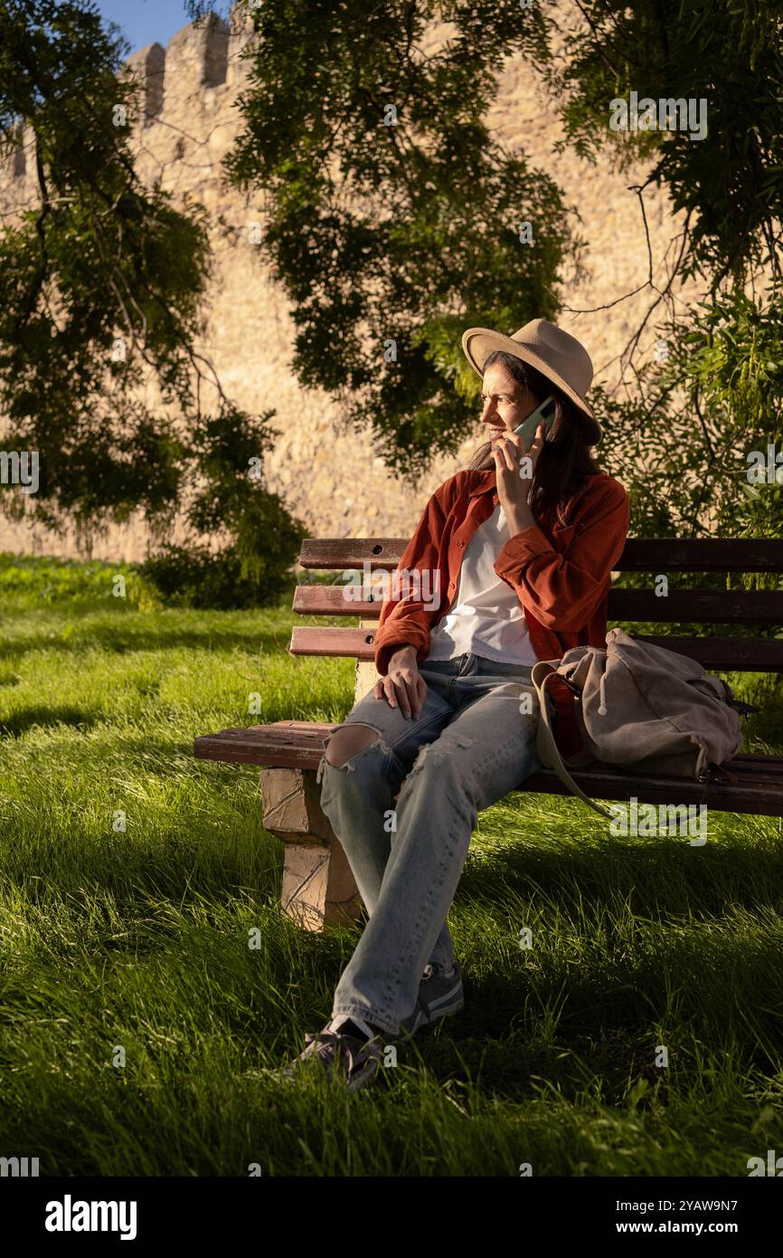 Femme touriste dans le grand chapeau se relaxant sur le banc dans le parc appréciant la conversation à l'aide du téléphone portable. Copier l'espace Banque D'Images