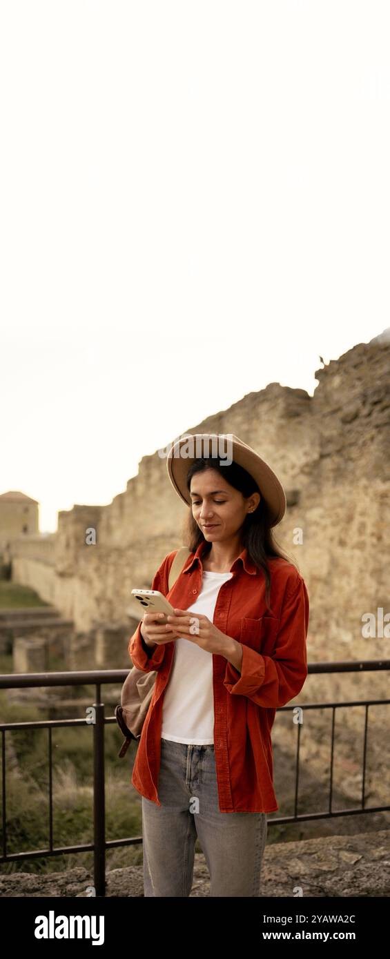 Une touriste vêtue d'un chapeau élégant se tient dans une ancienne forteresse, utilise un smartphone et dirige son blog de voyage. Bannière verticale Banque D'Images