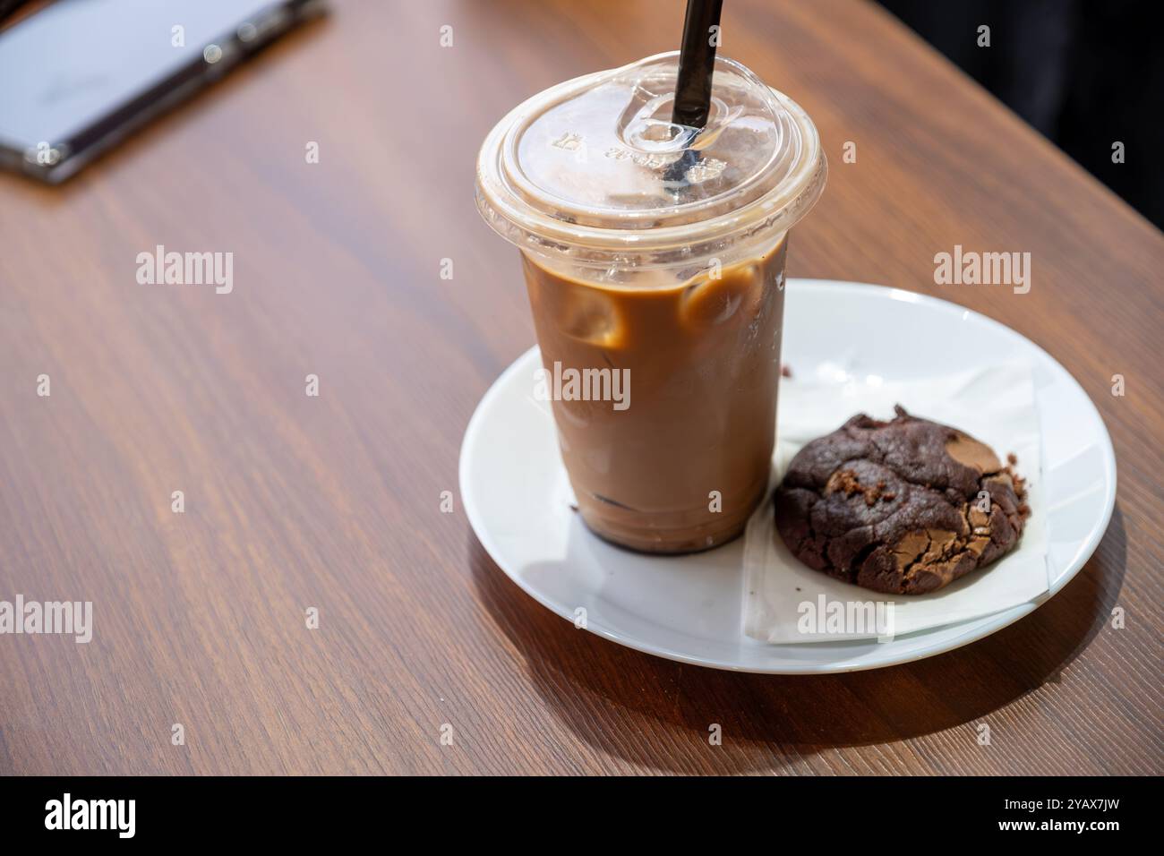 Café glacé réfrigéré servi dans une tasse en plastique avec une paille noire, accompagné d'un biscuit au chocolat sur une assiette blanche. Parfait pour une pause café confortable Banque D'Images