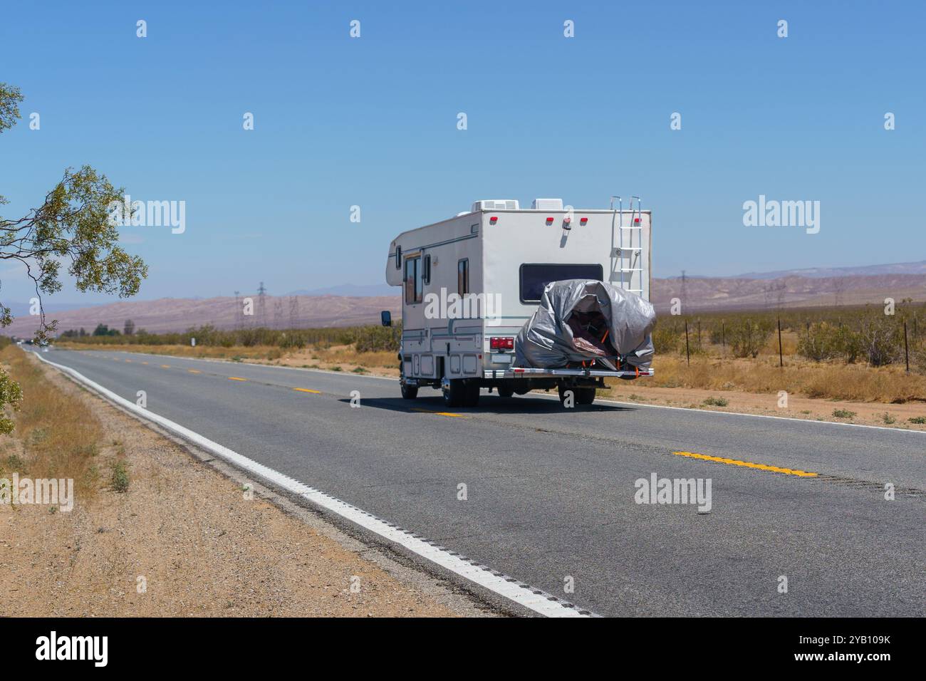 Un camping-car blanc avec une échelle et un véhicule couvert à l'arrière conduit sur une route pavée à travers un paysage désertique avec un ciel bleu clair. Banque D'Images