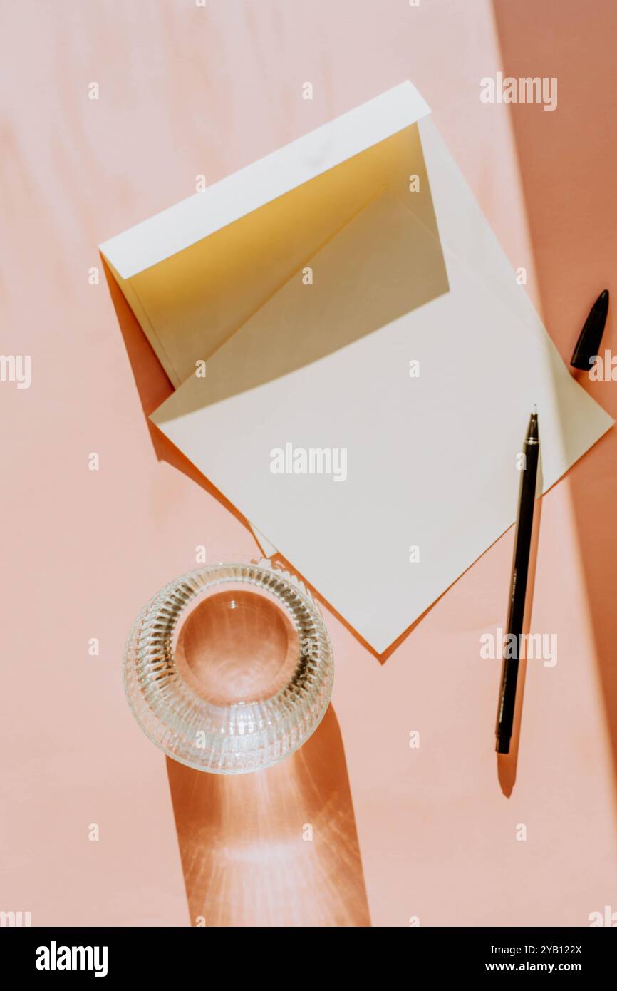 Ouvrez l'enveloppe et le papier blanc sur une surface douce de couleur pêche, accompagnés d'un stylo et d'un verre d'eau. L'éclairage chaud crée des ombres douces, addin Banque D'Images