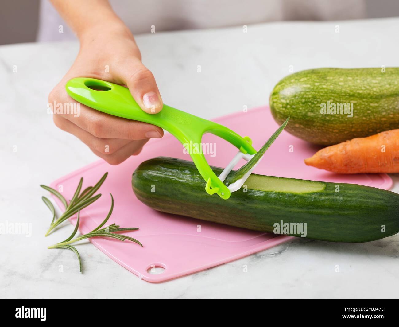 Un enfant épluche un concombre sur une planche à découper avec un éplucheur de légumes, gros plan d'une main tenant un outil pour nettoyer les légumes et les fruits Banque D'Images