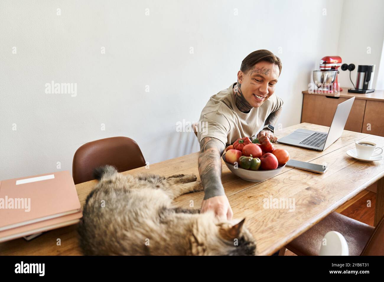 L'homme souriant interagit avec son chat tout en travaillant à une table en bois remplie de fruits frais. Banque D'Images