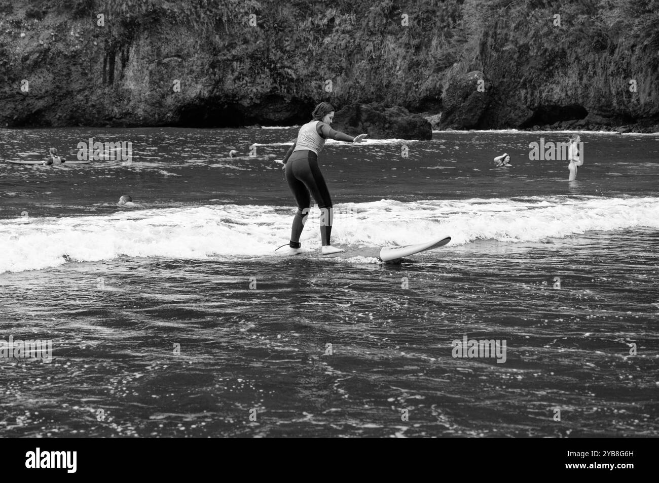 Une jeune surfeuse pratique son équilibre sur une petite vague lors d’une leçon de surf à la plage de Seixal, capturée en noir et blanc Banque D'Images