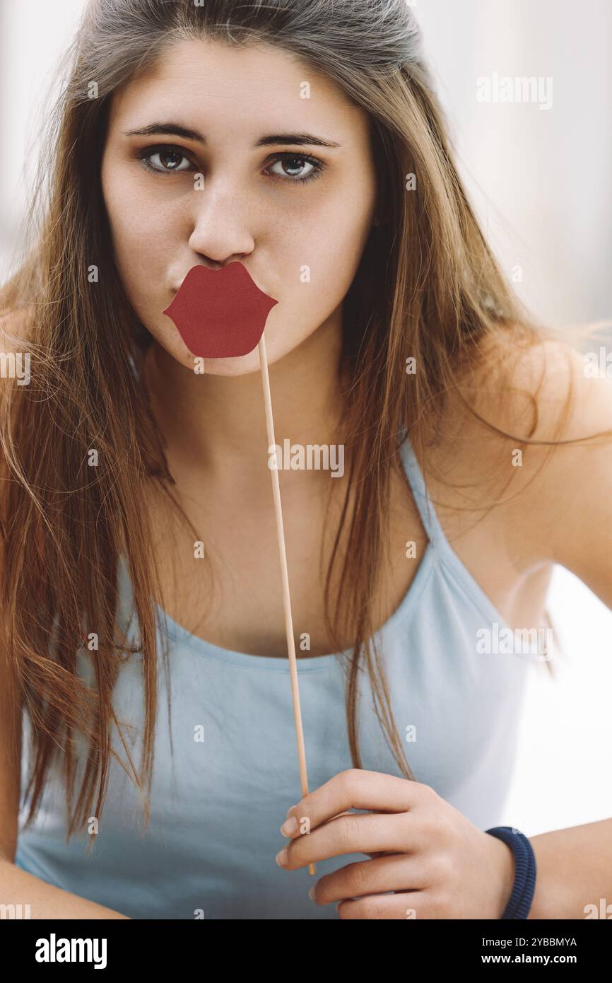 Jeune femme tient ludique un accessoire drôle de lèvres, exsudant la joie et l'excitation dans une cabine photo. Ses longs cheveux bruns descendent en cascade tandis qu'elle sourit au cheerfu Banque D'Images