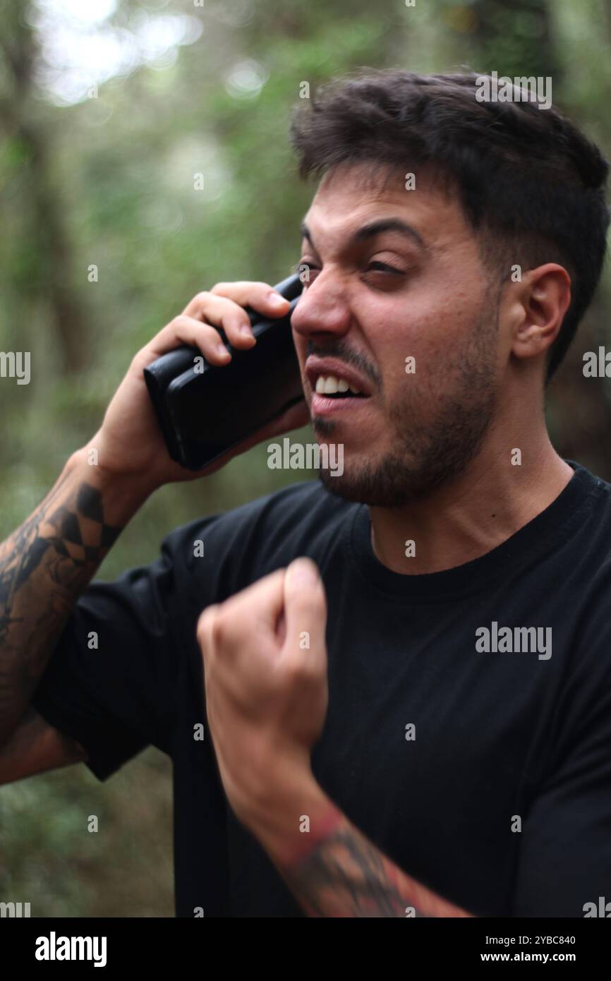 Un jeune homme frustré crie dans son téléphone portable pendant un appel téléphonique intense, debout au milieu d'une forêt paisible Banque D'Images