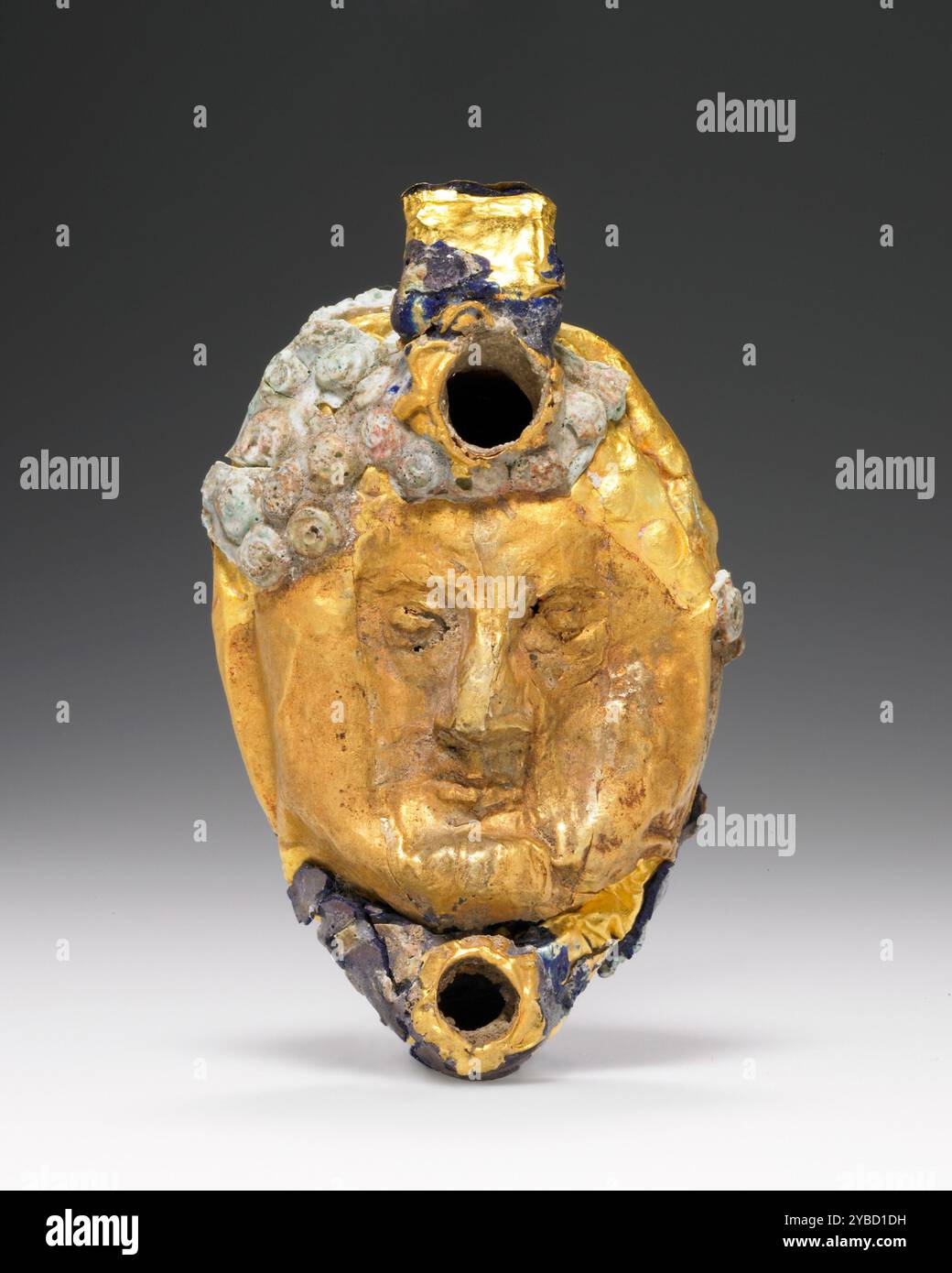 Lampe avec masque recouvert d'une feuille d'or, 1st Century B.C. - 1st Century A.D. informations supplémentaires : lampe miniature avec intérieur fait d'une substance indéterminée, sur laquelle une feuille d'or a été appliquée. L'image sur le dessus de la lampe apparaît en relief sur la feuille d'or comme une tête mâle, qui remplit la surface supérieure entre la poignée et la buse. Des ronds de verre vert pâle façonnés en petits cercles concentriques ont été appliqués en imitation de boucles de cheveux. Serrures principalement vert pâle, parfois tendant vers le rose. La poignée de sangle est recouverte de verre doré et bleu. Trou de remplissage près de la poignée entouré par le bord de sm Banque D'Images