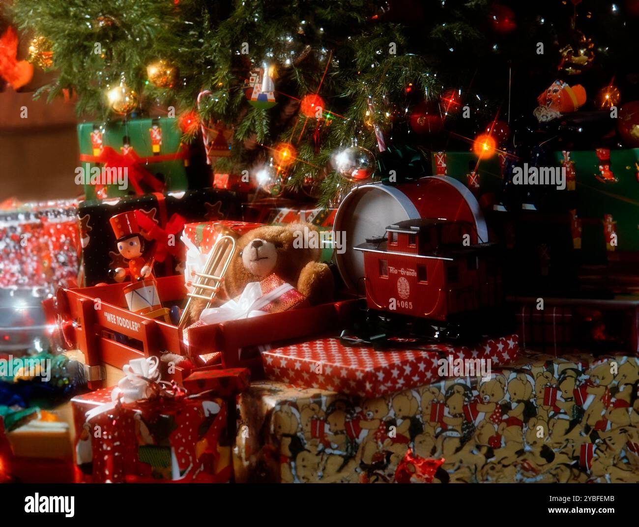 Une collection de cadeaux emballés se trouve sous un sapin de Noël décoré d'ornements et de lumières. Un train de jouets rouge, un ours en peluche avec une trompette, et un Banque D'Images