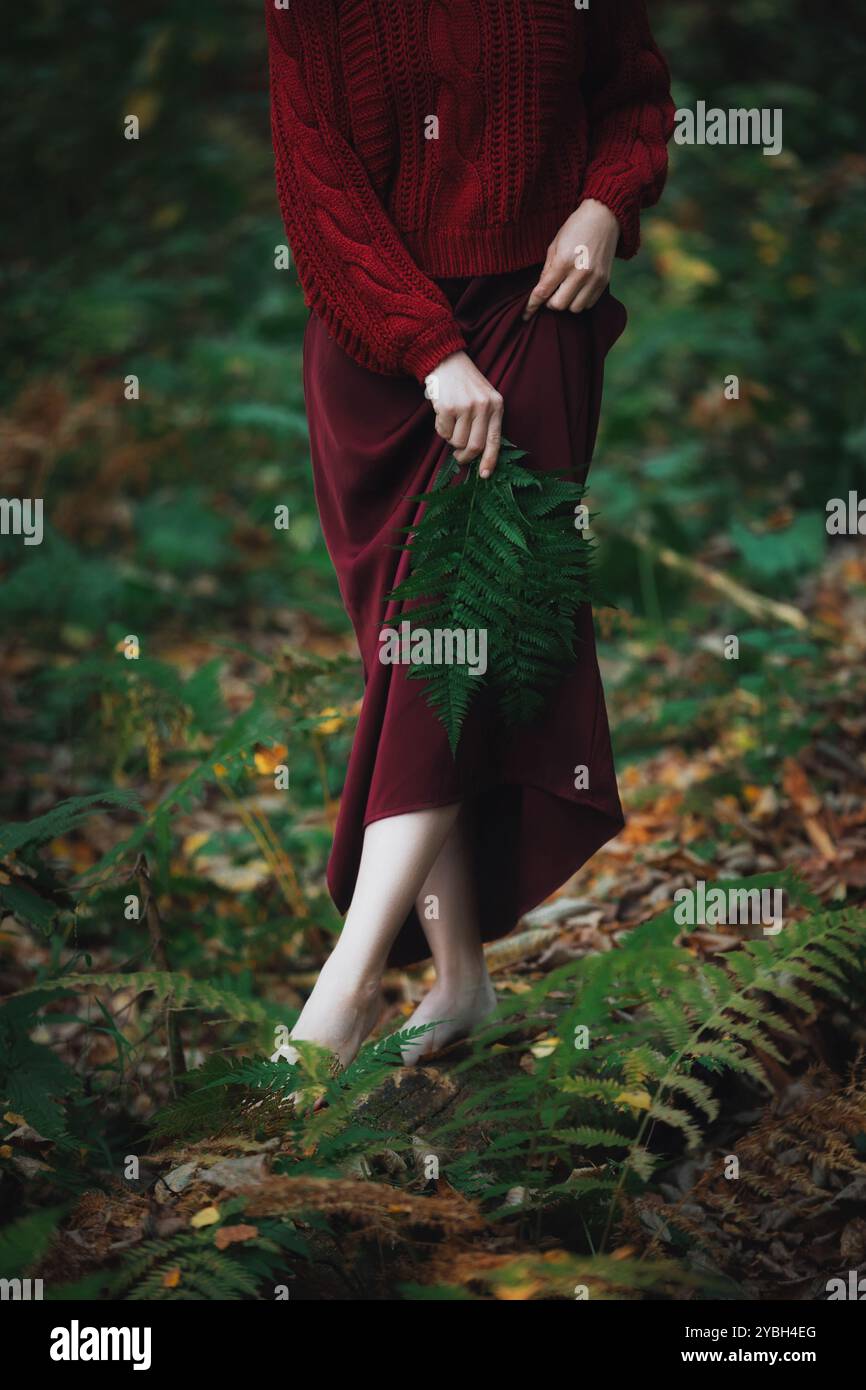 Une femme portant une robe Bordeaux se tient pieds nus dans un cadre de forêt sereine, entourée de fougères vertes vibrantes. Elle tient doucement une grande feuille, embodyi Banque D'Images
