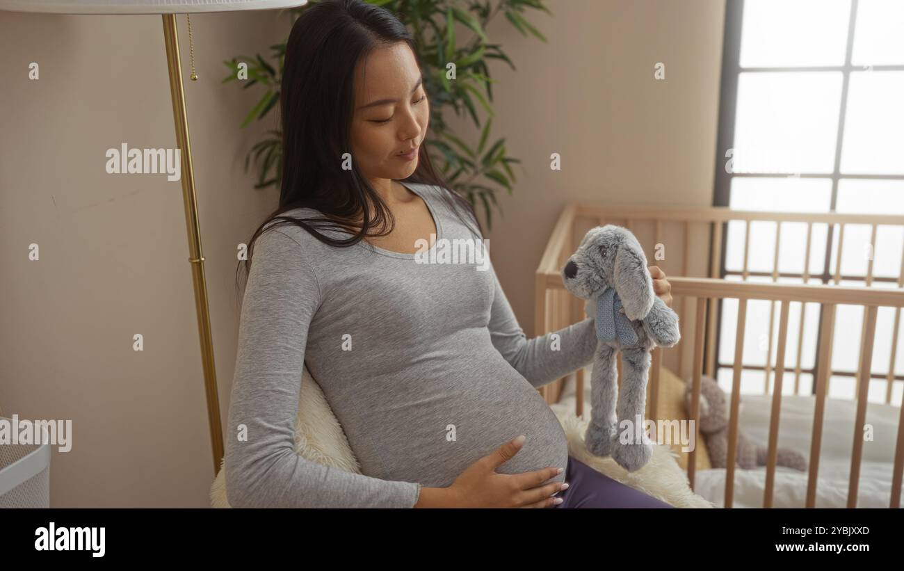 Femme enceinte assise sur un lit dans une chambre confortable tenant un animal en peluche à côté d'un berceau de bébé Banque D'Images