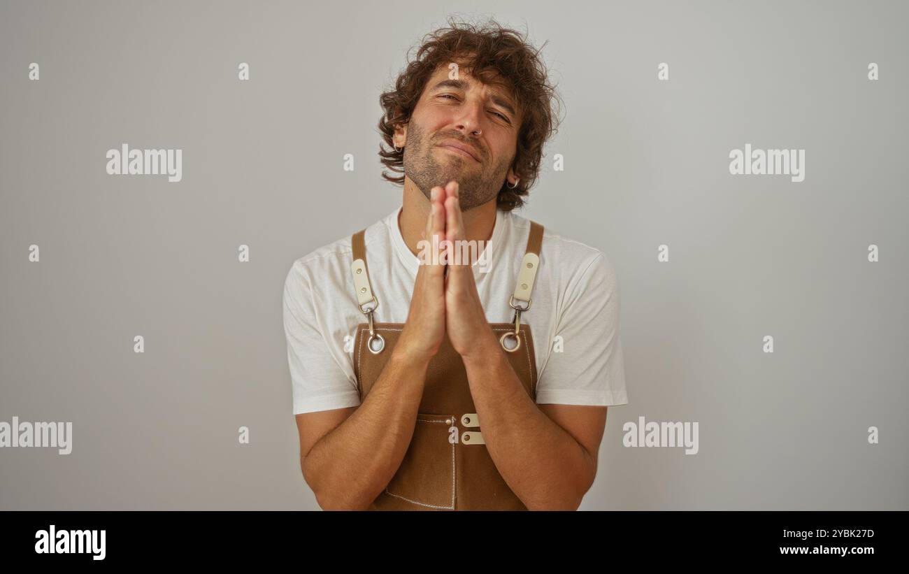 Jeune homme aux cheveux bouclés portant une combinaison brune et une chemise blanche pose avec les mains ensemble dans un geste de plaidoyer sur un fond blanc isolé Banque D'Images