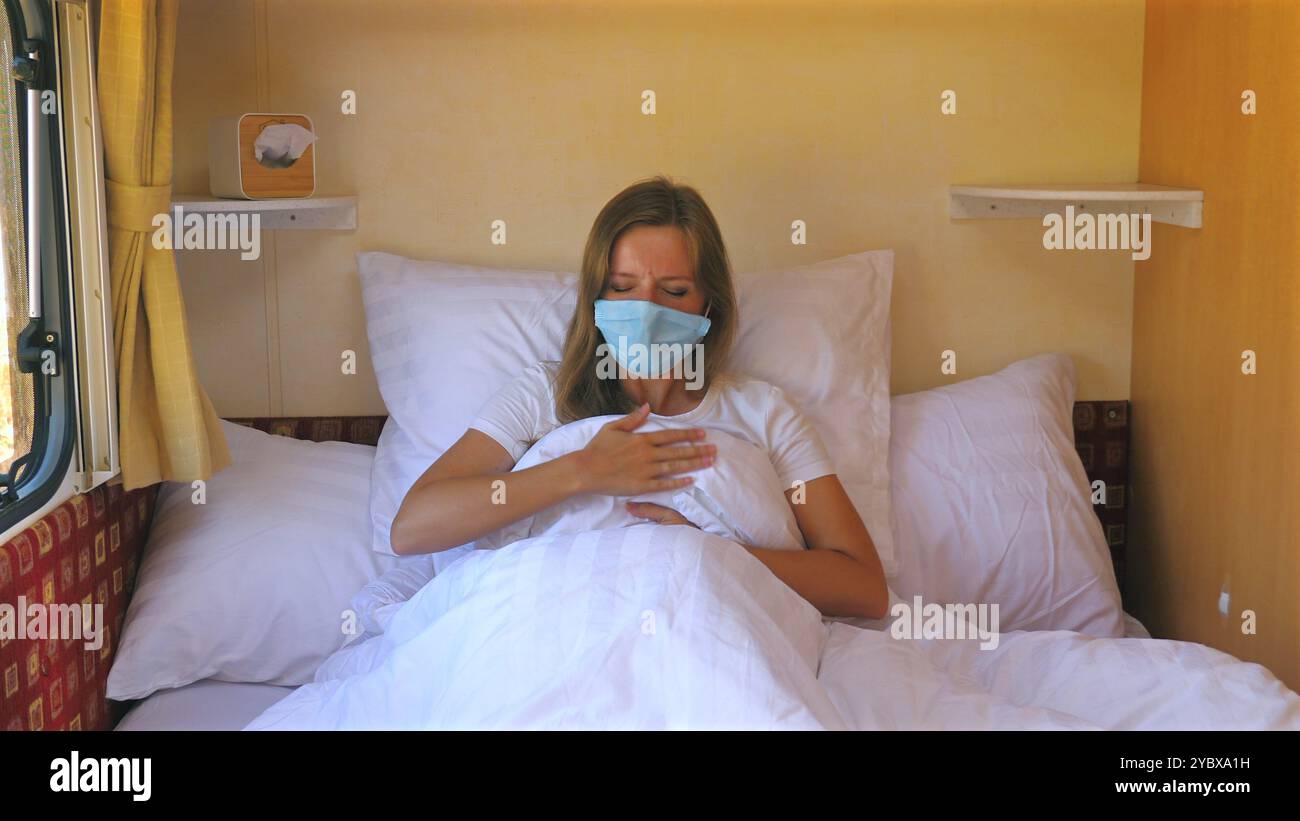 Femme portant un masque facial, assise sur le lit et toussant, à l'intérieur d'une maison confortable Banque D'Images
