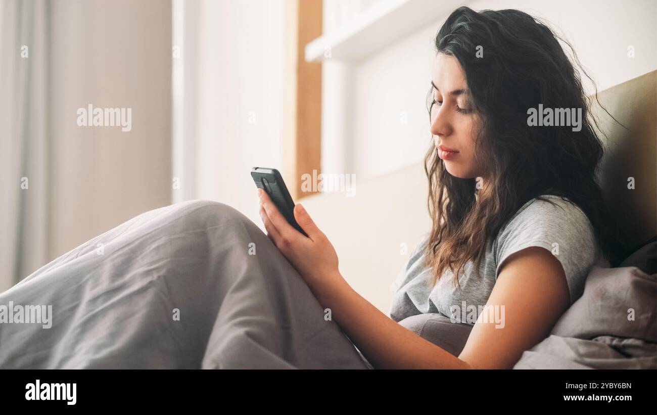 Mauvaise nouvelle. Femme frustrée. Chat mobile. Message de femme déçue bouleversée sur le smartphone assis au lit dans la chambre du matin claire. Banque D'Images