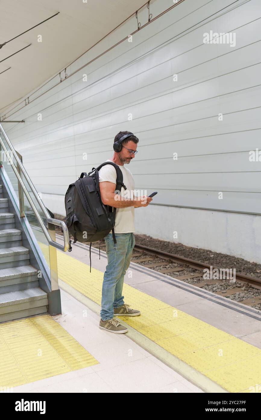 Homme d'âge moyen dans des vêtements décontractés avec sac à dos en utilisant smartphone pour E-ticket en attendant le train à la gare Banque D'Images