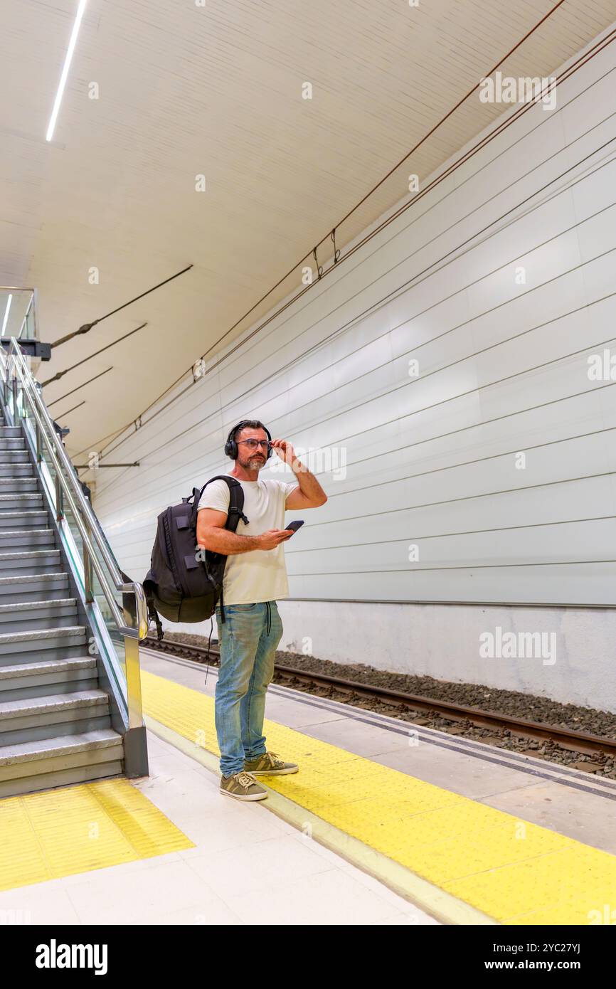 Homme d'âge moyen dans des vêtements décontractés avec sac à dos en utilisant smartphone pour E-ticket en attendant le train à la gare Banque D'Images