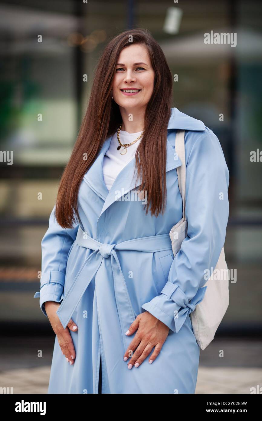Portrait charmante grande taille femme avec de longs cheveux sourient en toute confiance à l'extérieur, porte un trench bleu clair à la mode et porte un sac en toile, showcas Banque D'Images