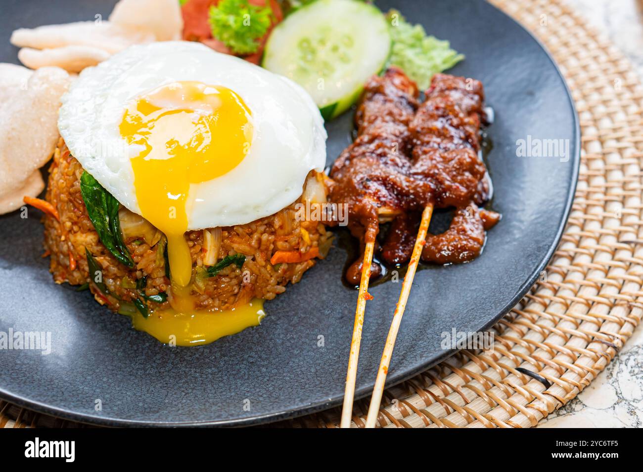 Une assiette de riz frit garnie d'un œuf ensoleillé, accompagné de brochettes satay, de concombres tranchés, de laitue et de tomates. Le plat est servi sur un bl Banque D'Images