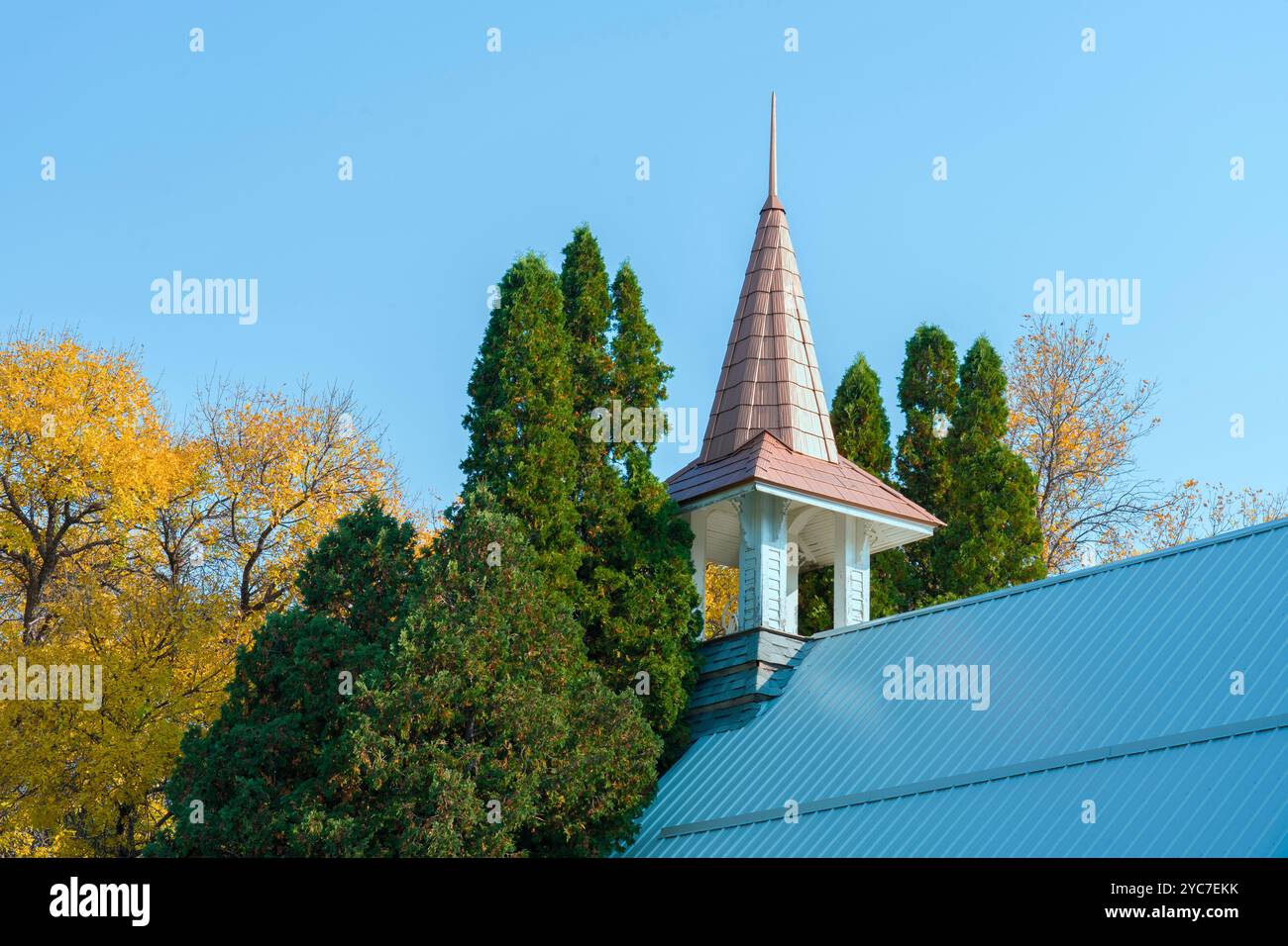 Vieux clocher de toit d'église montre contre un ciel bleu avec des cimes d'arbres environnants de pins et d'autres arbres d'automne avec des feuilles jaunes tombantes. Banque D'Images