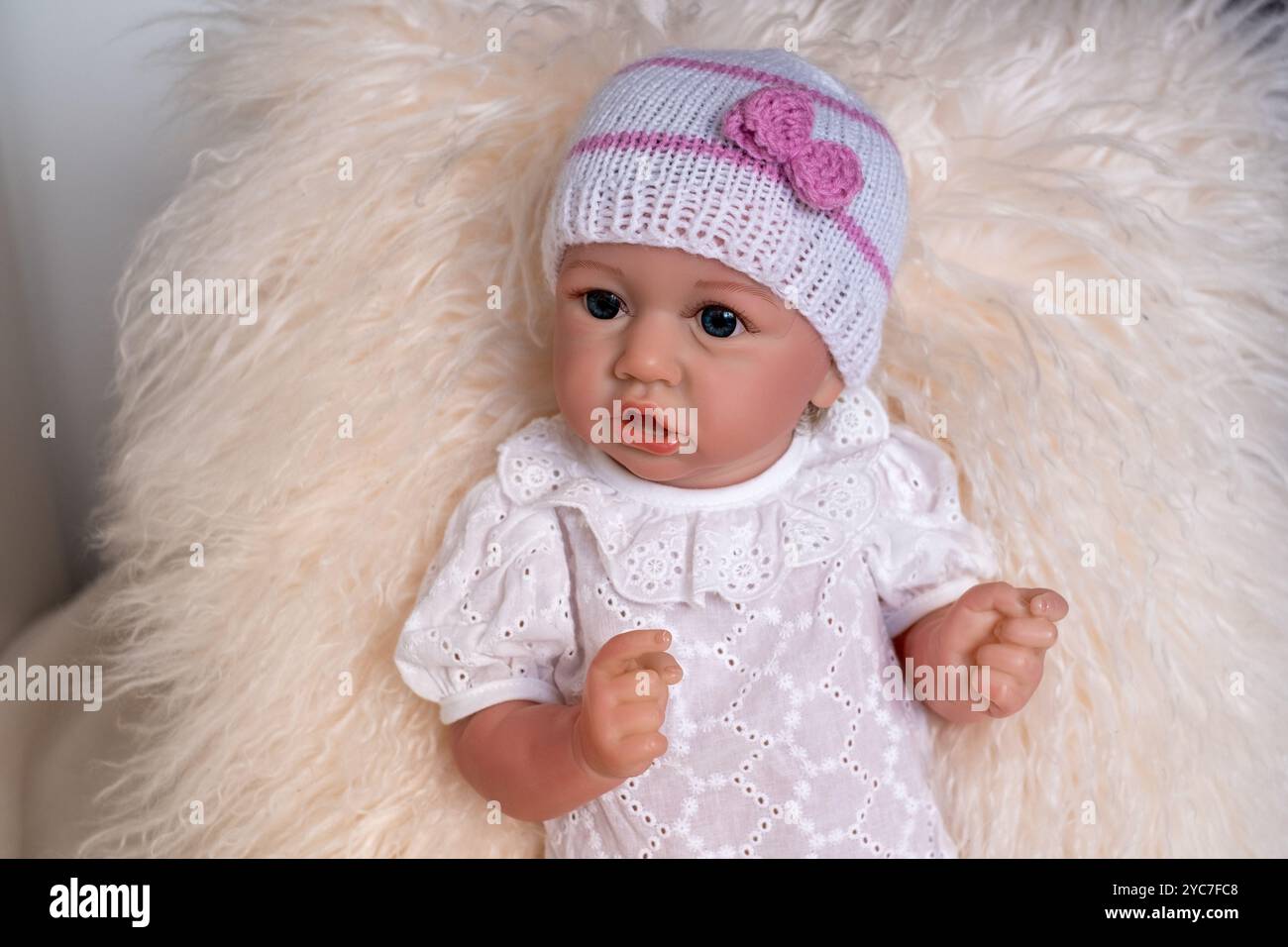 Une poupée bébé réaliste repose sur une couverture douce et moelleuse, vêtue d'une délicate tenue blanche et d'un bonnet tricoté aux accents roses, dégageant charme et tendresse Banque D'Images