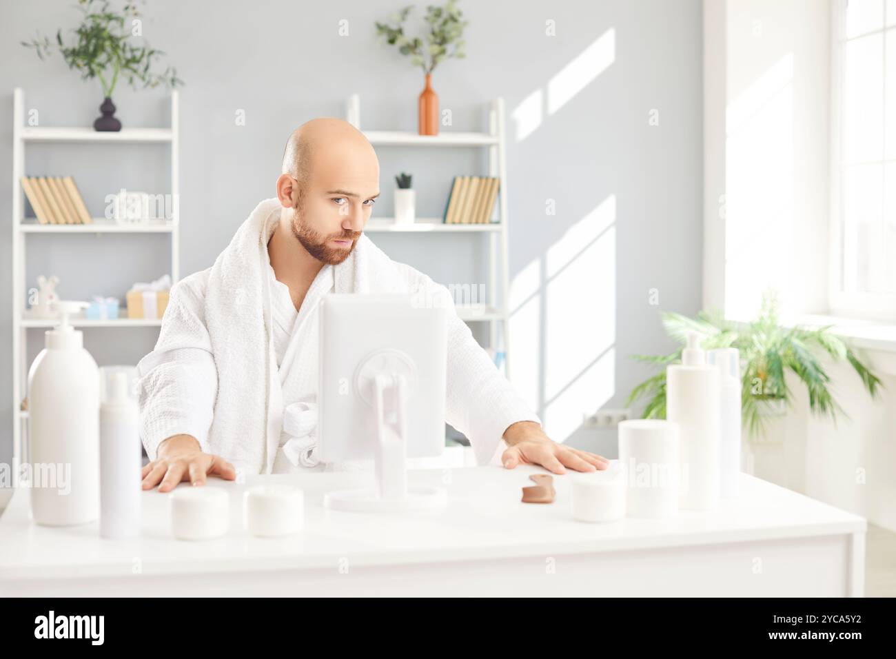 Homme drôle en peignoir blanc assis à table avec des bouteilles cosmétiques ayant la journée de beauté à la maison. Banque D'Images