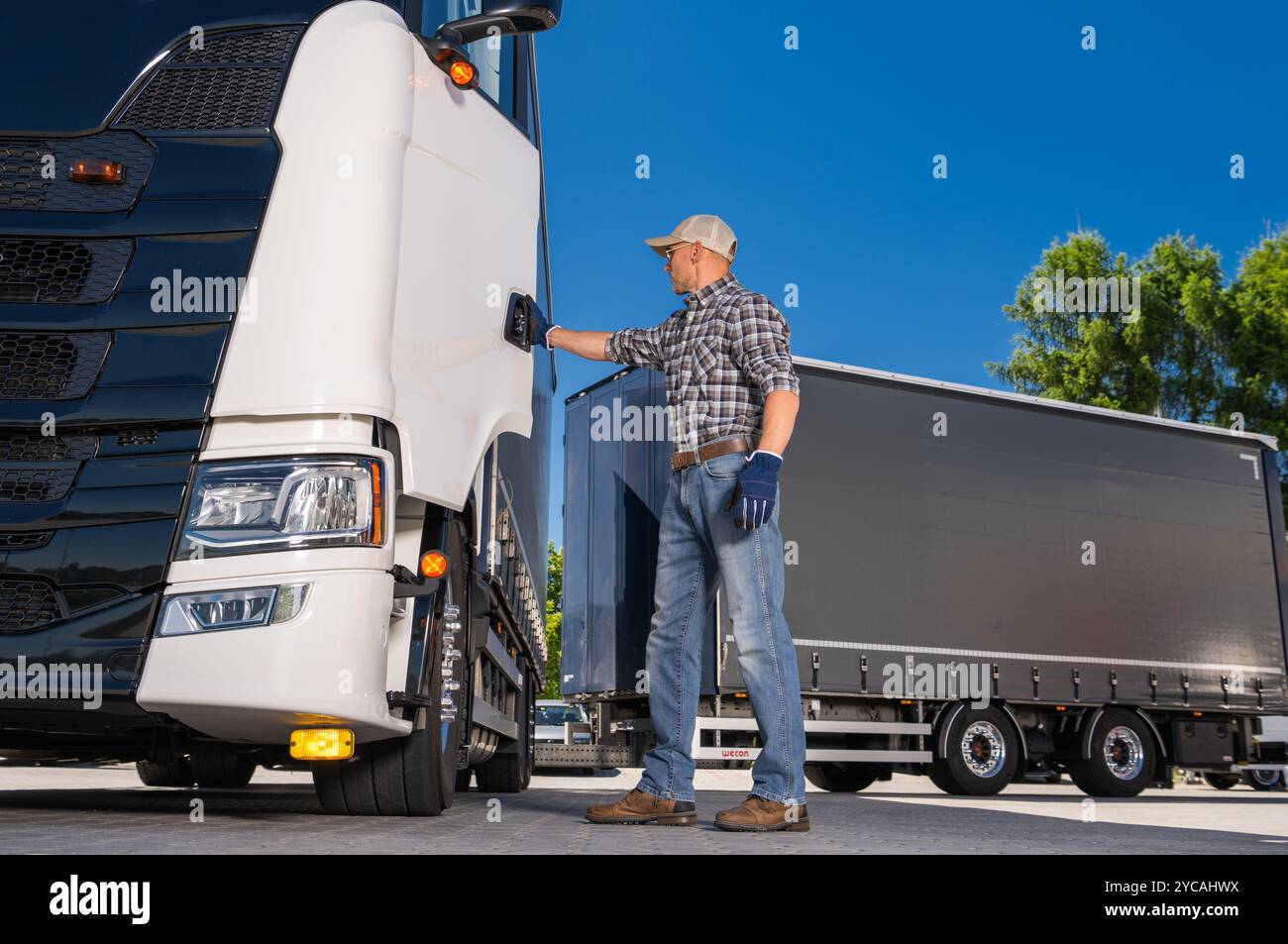 Un conducteur de camion, portant des gants et un chapeau, vérifie le réservoir de carburant d'un gros camion stationné sur un quai de chargement sous ciel dégagé. Banque D'Images