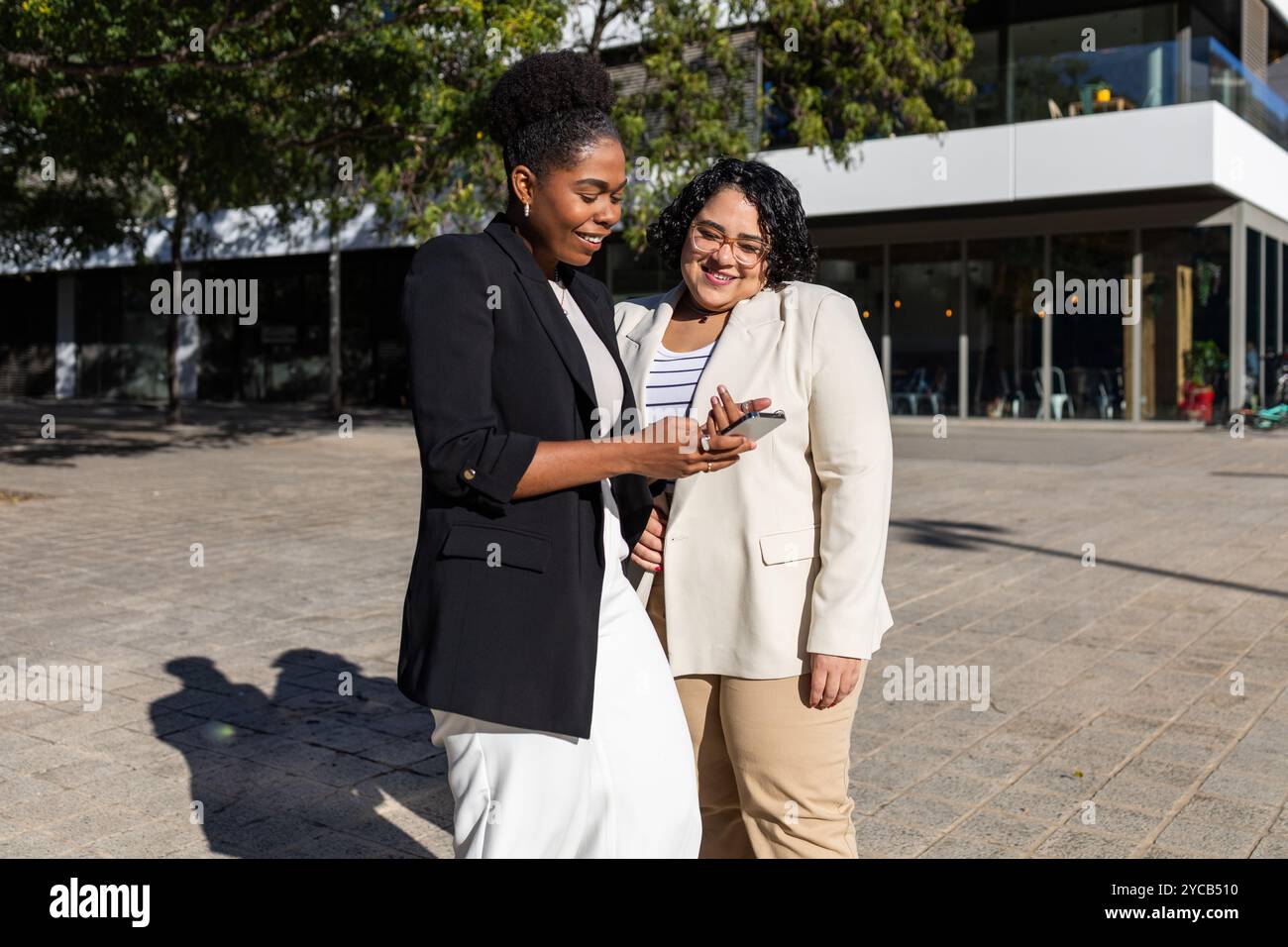 Deux femmes d'affaires, une afro-américaine et une hispanique, utilisant un smartphone ensemble tout en se tenant à l'extérieur. Les deux sourient et regardent le scr Banque D'Images