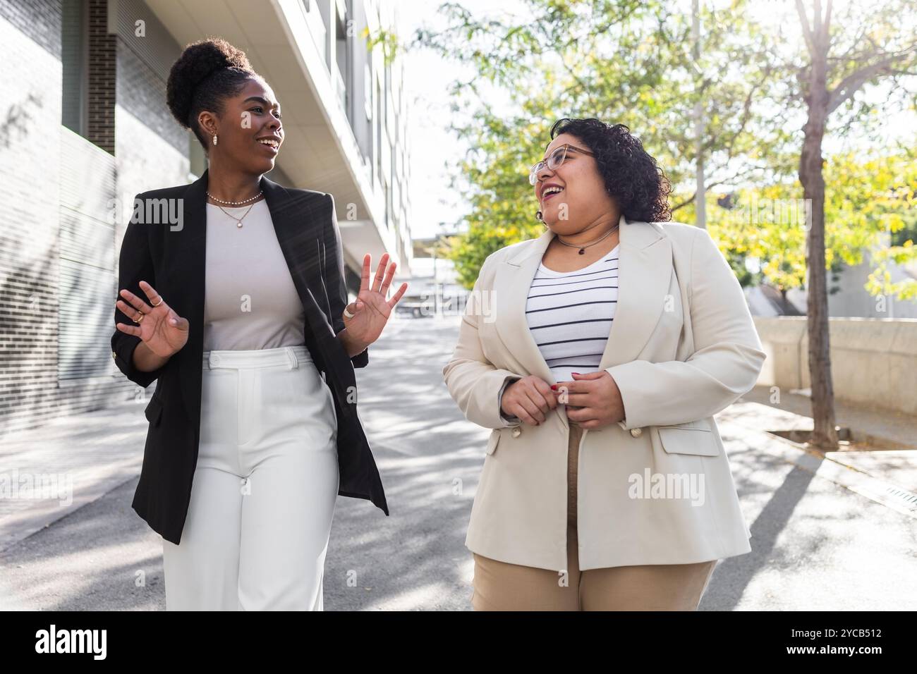 Deux femmes professionnelles appréciant une conversation tout en marchant dans une rue ensoleillée de la ville. La femme afro-américaine fait des gestes animés comme les deux rires toge Banque D'Images