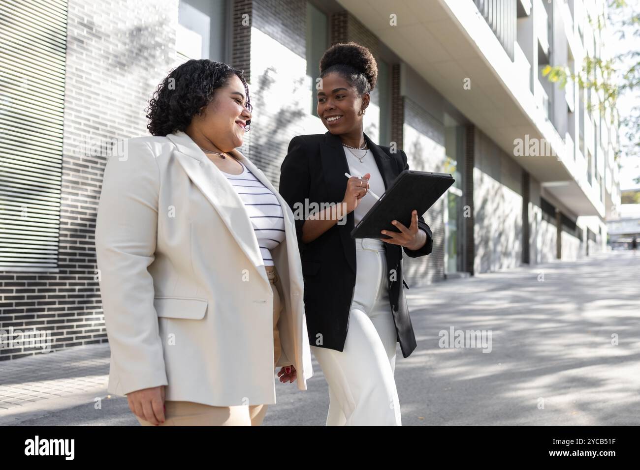 Deux femmes d’affaires afro-américaines s’engagent dans une discussion tout en marchant à l’extérieur, l’une tenant une tablette numérique. Les deux sont souriants et professionnellement dr. Banque D'Images